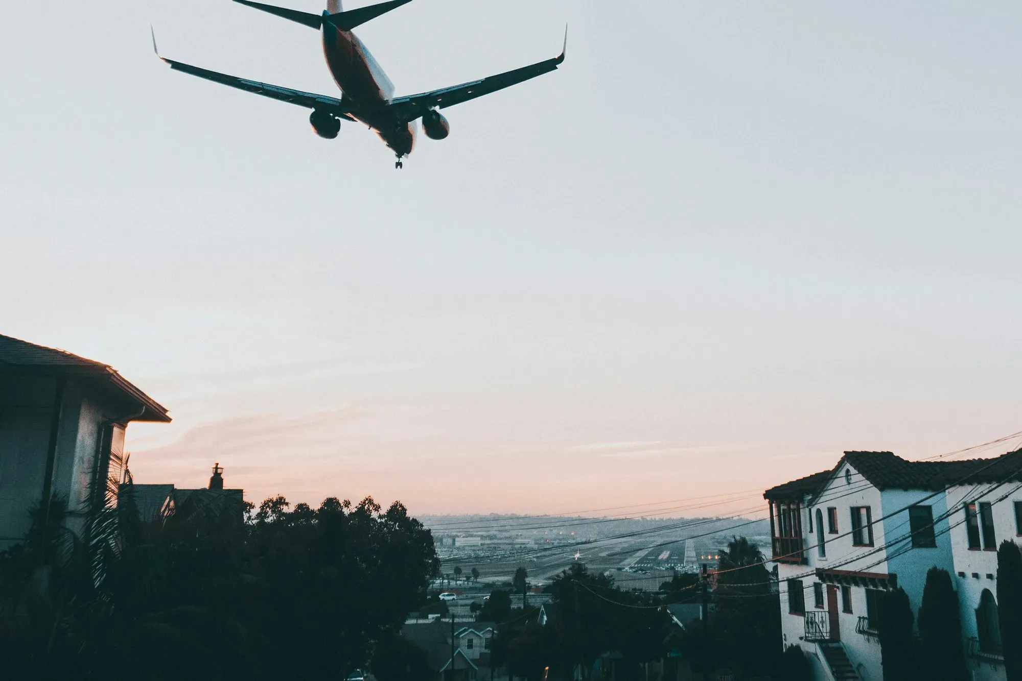 A jet on final approach passes low over Bankers Hill's rooftops at dusk, the airport runway visible in the distance below