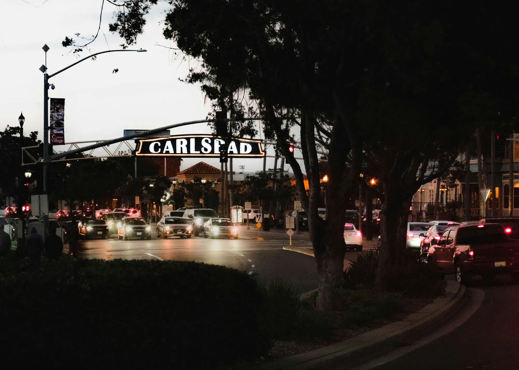 The lit overhead Carlsbad sign above the Village at dusk, traffic passing underneath