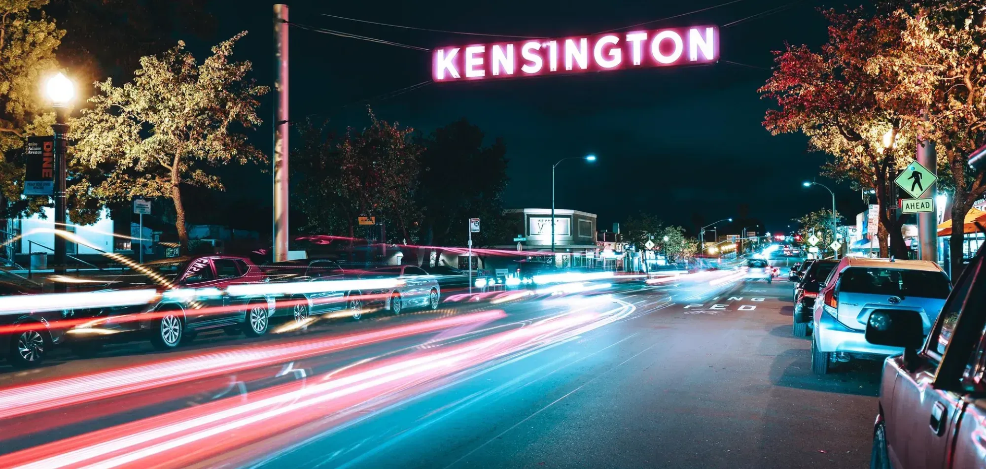 The lit Kensington sign over Adams Avenue at night, traffic streaking below it