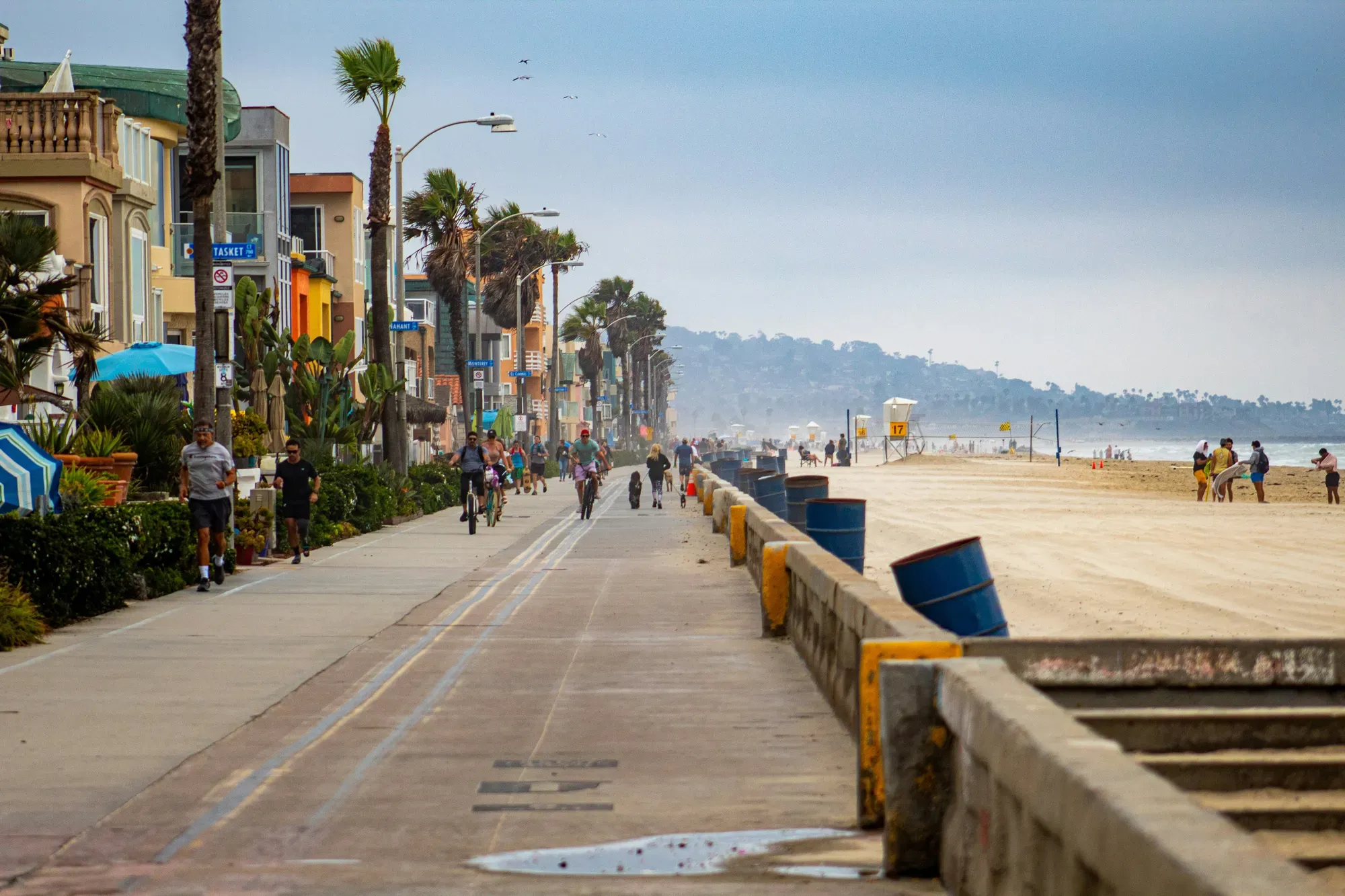 Joggers, cyclists, and dog walkers on the Mission Beach boardwalk on a hazy morning, the coast curving north toward Pacific Beach