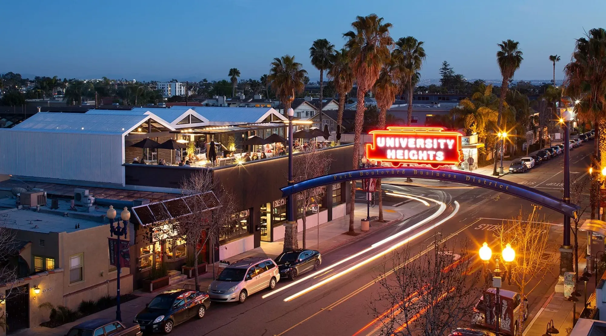 The lit University Heights sign arching over Park Boulevard at night, light trails from passing cars below
